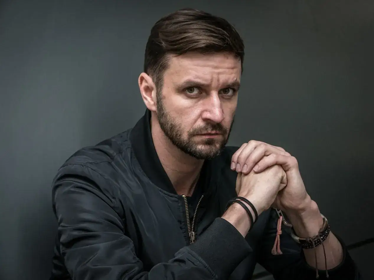 Mężczyzna z zarostem w czarnej kurtce, z dłońmi złożonymi przed sobą. Może to być kadr z filmu 