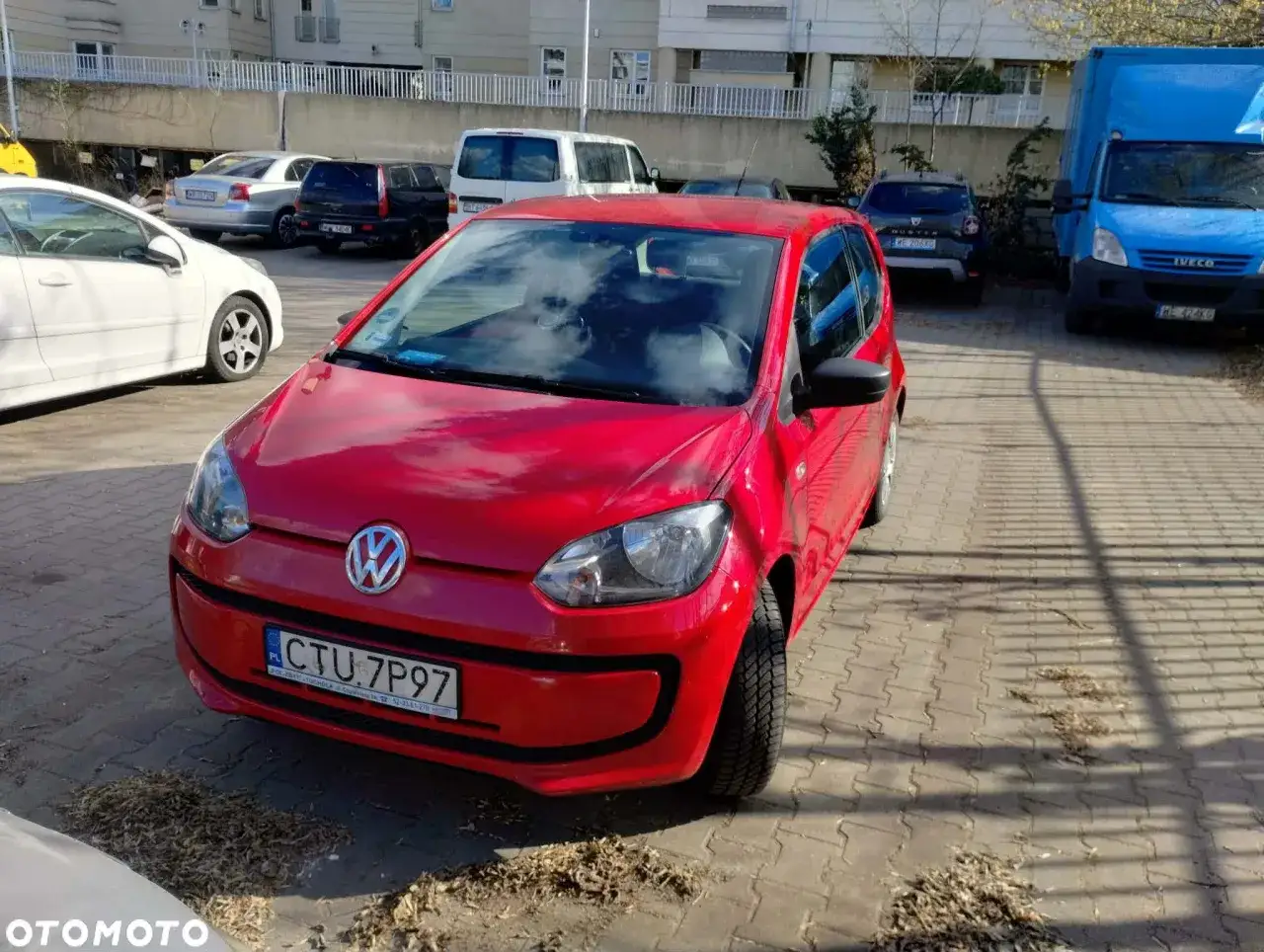 Czerwony Volkswagen Up stoi na parkingu obok innych samochodów.