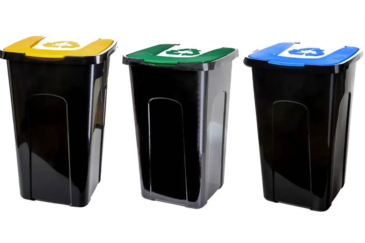 Trzy czarne kosze na śmieci z kolorowymi pokrywami: ż&oacute;łtą, zieloną i niebieską. Każda pokrywa ma symbol recyklingu. To niebieski pojemnik na śmieci, do kt&oacute;rego wrzucamy papier.