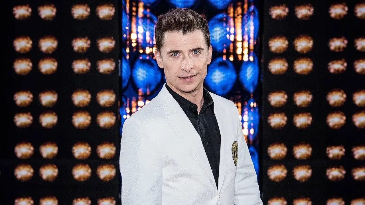 Kacper Kuszewski w białej marynarce, z żoną u boku, pozuje na tle błyszczącej sceny.