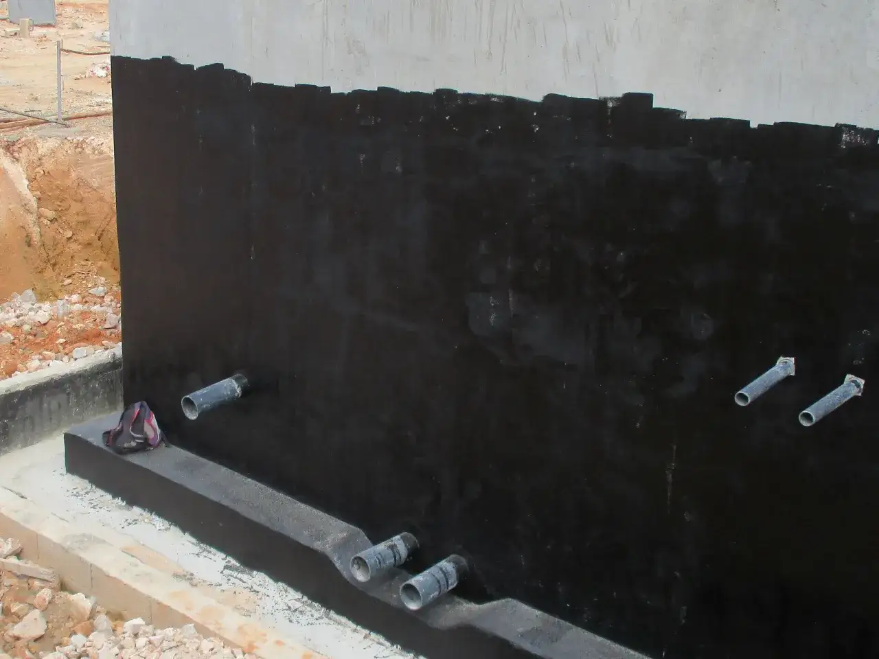 Application d'une membrane noire pour l'&eacute;tanch&eacute;it&eacute; d'un mur enterr&eacute;. Des tuyaux sortent du mur, pr&ecirc;ts pour le drainage.