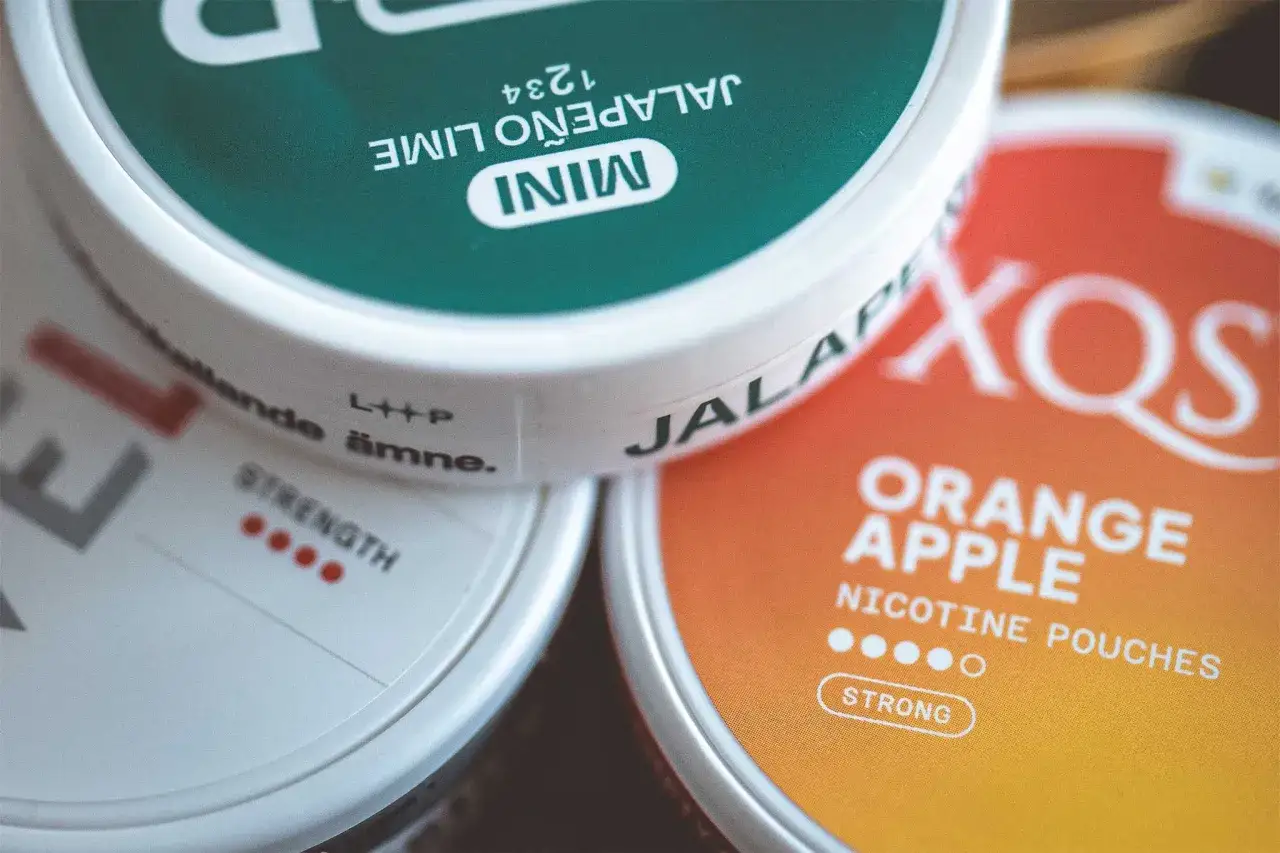 Tre dosor nikotinpåsar: en grön med jalapeño lime, en vit med styrkeindikator, och en orange med orange äpple. En snus styrka tabell kan jämföras med dessa.