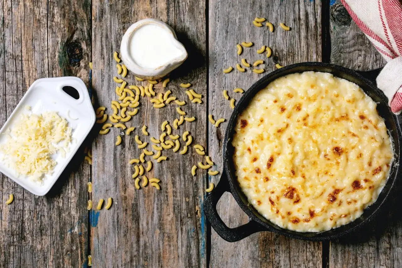 Mac and cheese oryginalny przepis: zapieczony makaron z serem w żeliwnej patelni, obok surowy makaron, starty ser i śmietanka.