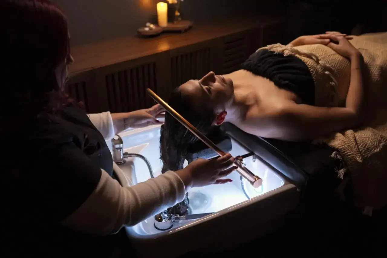 Relaks w salonie piękności. Kobieta leży na fotelu, a stylistka wykonuje zabieg head spa, delikatnie masując jej włosy.