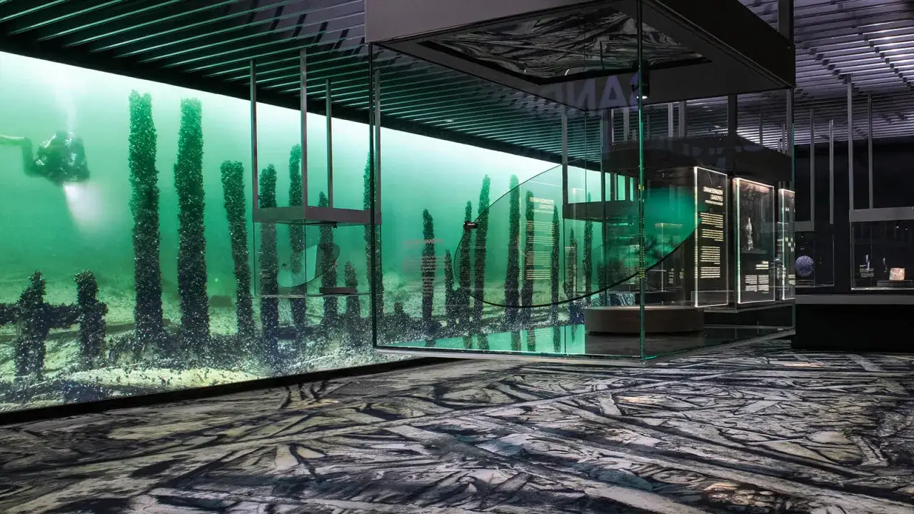 En dykare utforskar en undervattensv&auml;rld med gamla tr&auml;p&aring;lar, en del av ett museum of wrecks. Glasmontrar visar artefakter fr&aring;n djupet.