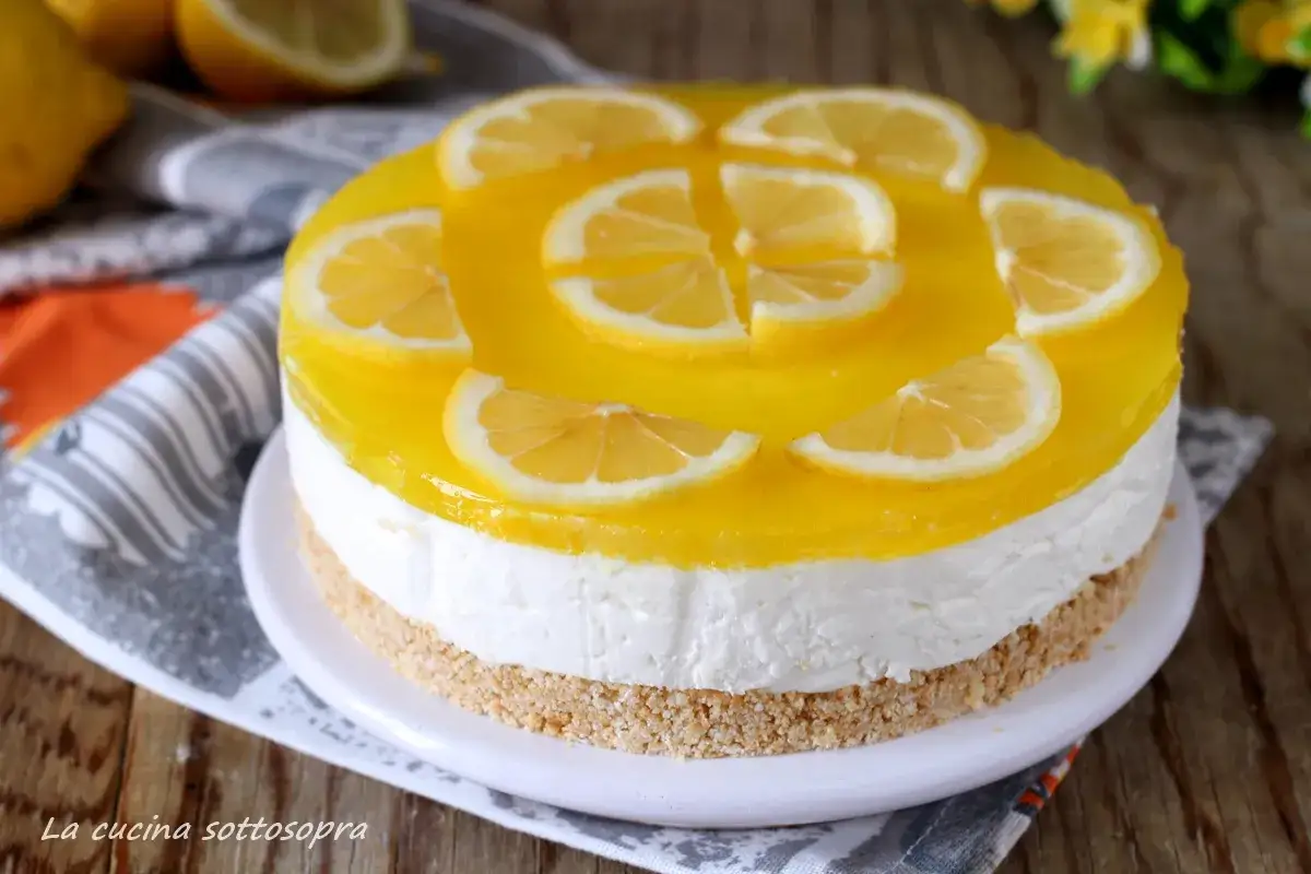 Torta fredda con base croccante, strato bianco e copertura gialla lucida, decorata con fette di limone. Una deliziosa mousse al limone pronta da gustare.