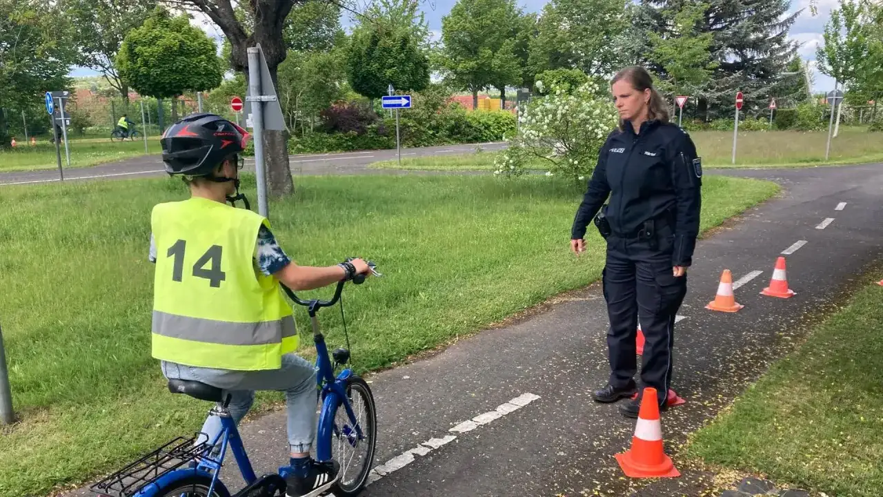 Polizistin leitet Fahrrad-Ausbildung f&uuml;r Kind mit Warnweste und Helm auf &Uuml;bungsstrecke mit H&uuml;tchen.