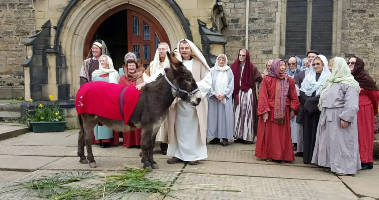 A donkey, adorned in a red blanket, stands before a group dressed for a Palm Sunday homily.