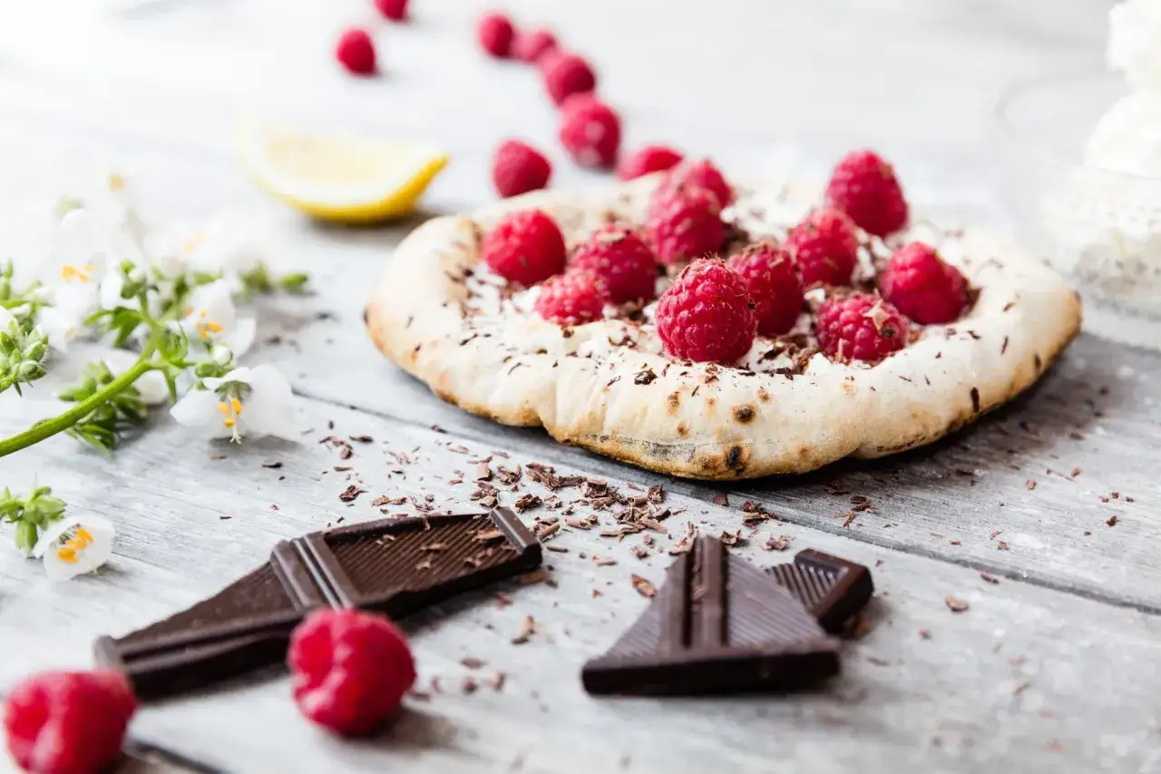Eine k&ouml;stliche Cookie Pizza mit frischen Himbeeren, Schokoladenraspeln und einem Zitronenschnitz auf Holztisch.