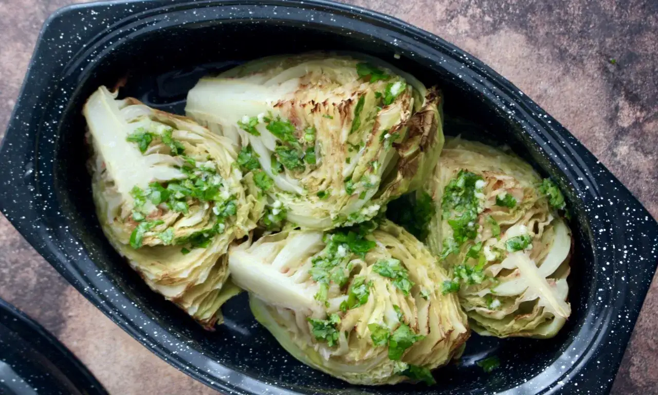 Quatre quartiers de choux verts au four, arrosés d'une sauce aux herbes et à l'ail, dans un plat noir émaillé.