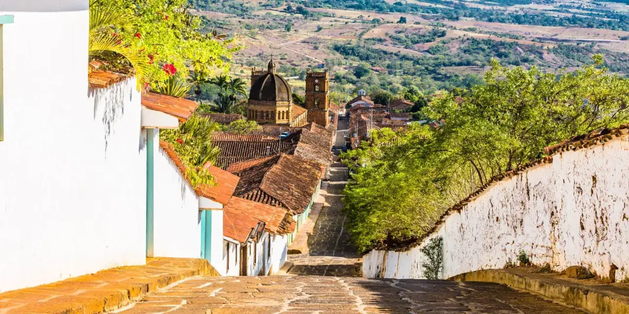 Calle empedrada desciende hacia un pueblo colonial con tejados rojos y una iglesia imponente. Un paisaje típico de los pueblos de Colombia.