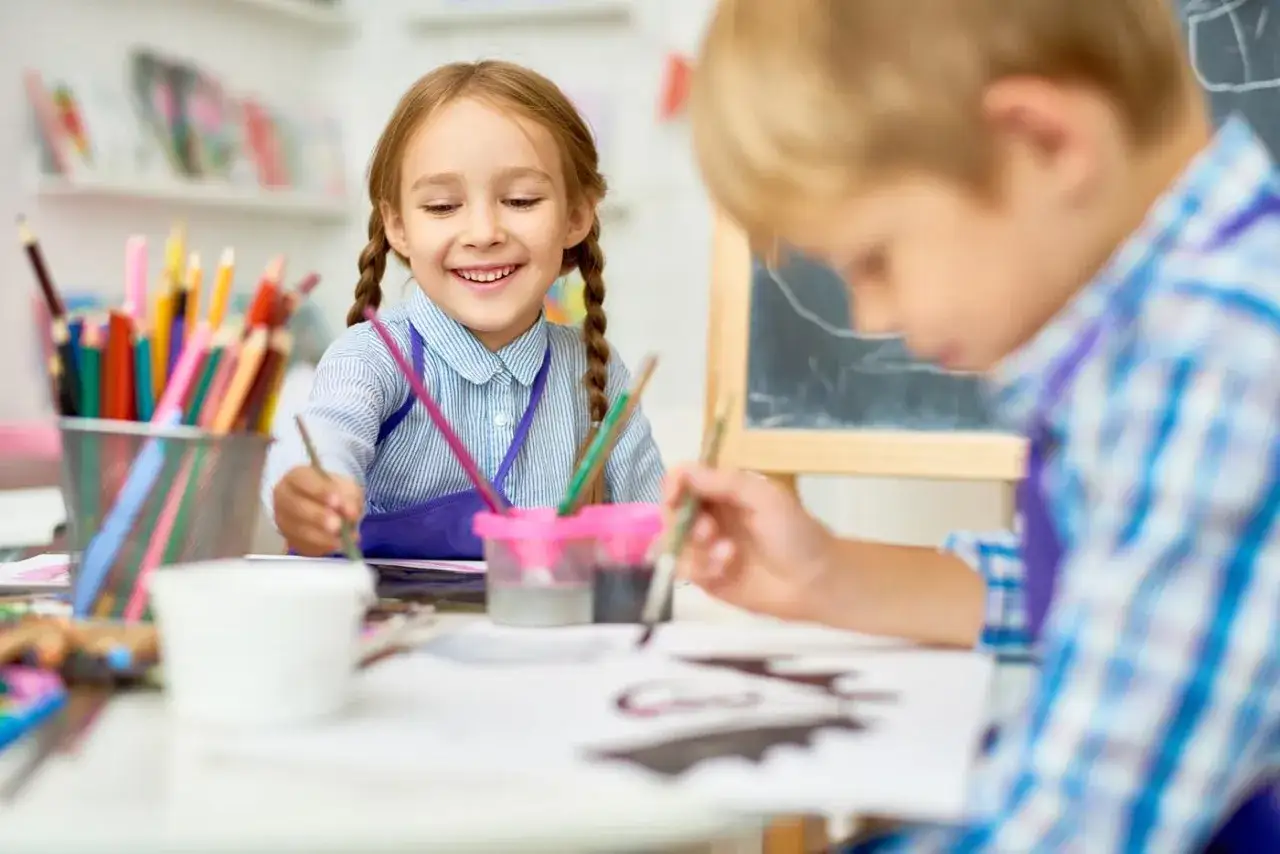 Fröhliche Kinder beim Malen im **Förderunterricht Grundschule**. Buntstifte und Farben liegen bereit.