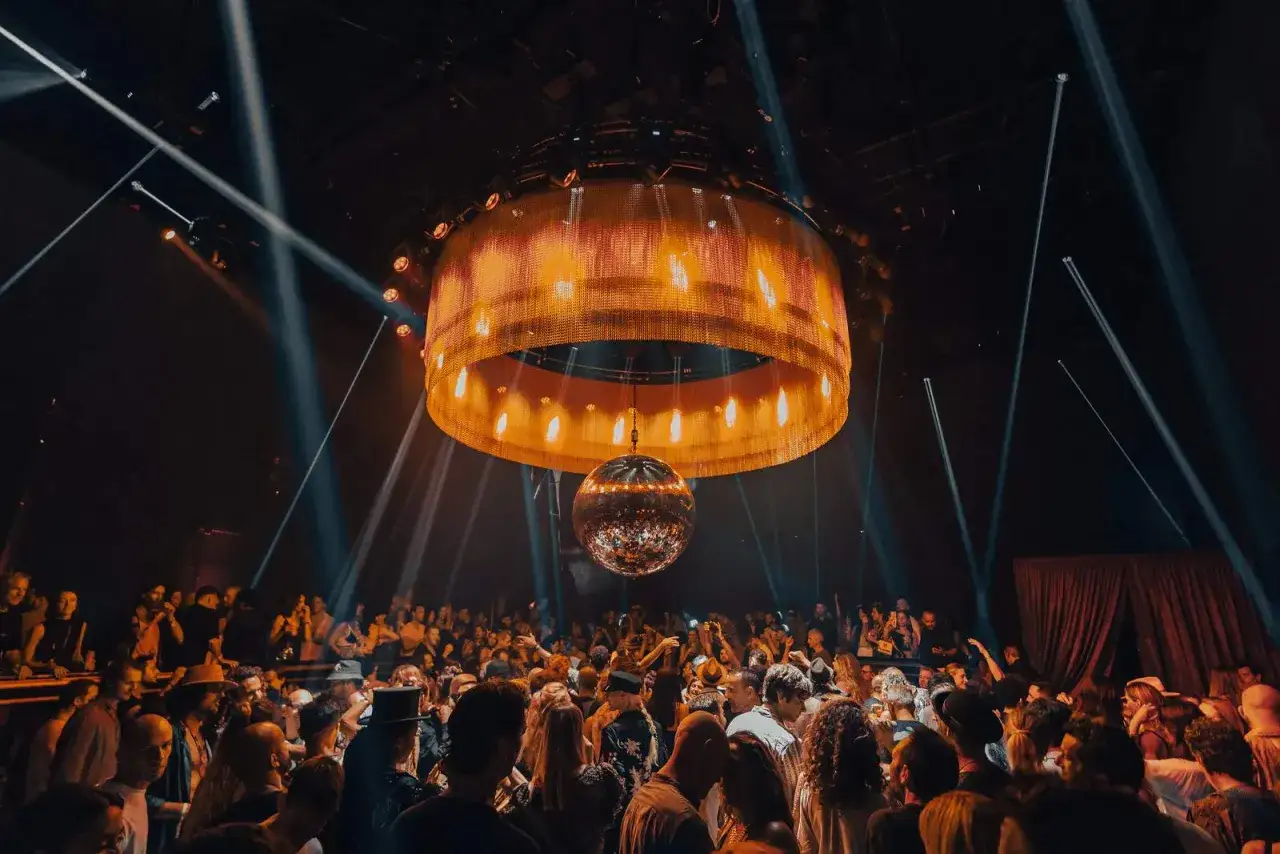 Folla in delirio sotto la maestosa lampada e la sfera da discoteca di IMS Ibiza. Luci e musica creano un'atmosfera elettrizzante.