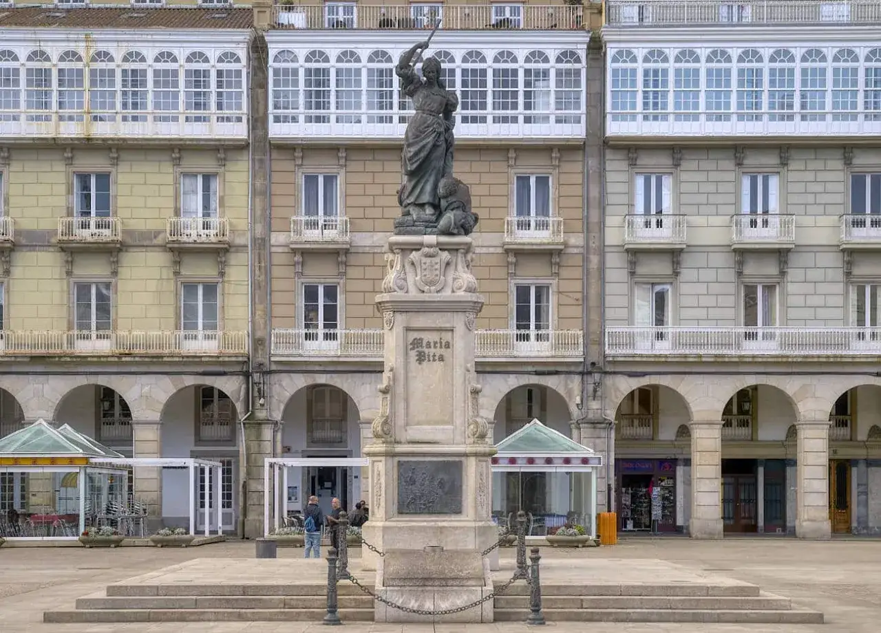 Monumento a María Pita en la plaza homónima, con edificios de balcones blancos y arcos.