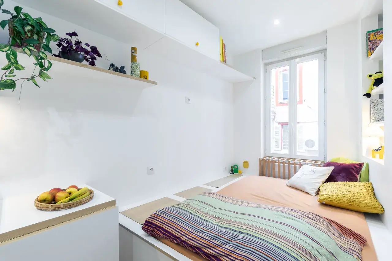 Chambre avec lit, coussins colorés, plantes et fruits. La **position bz** du lit est optimisée pour l'espace.