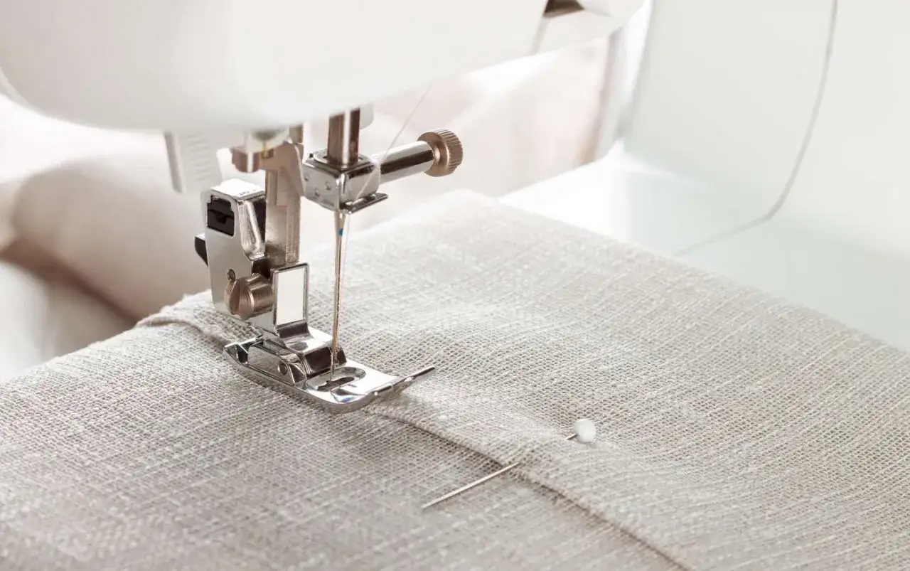 A sewing machine's topstitching foot guides a needle through beige fabric, creating a neat seam.