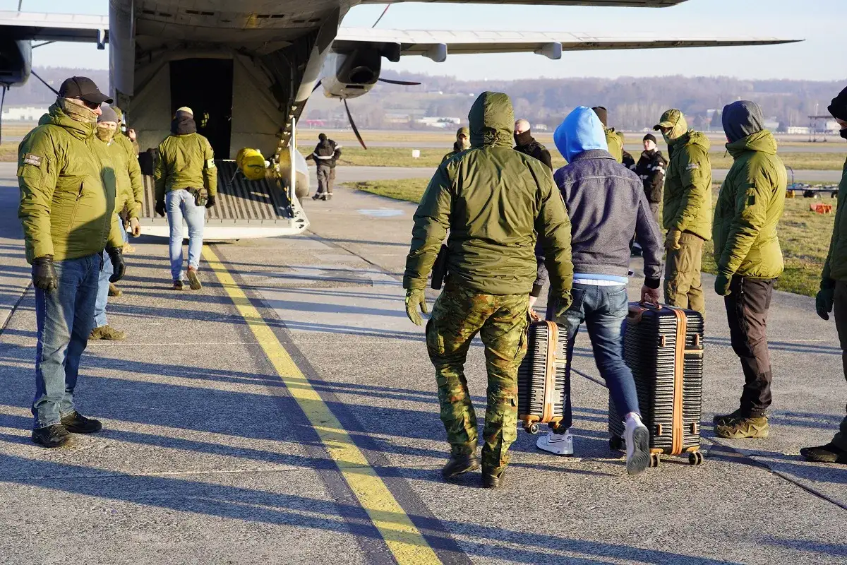 Osoby z bagażami zmierzają do samolotu. Może to być początek podróży lub deportacja.