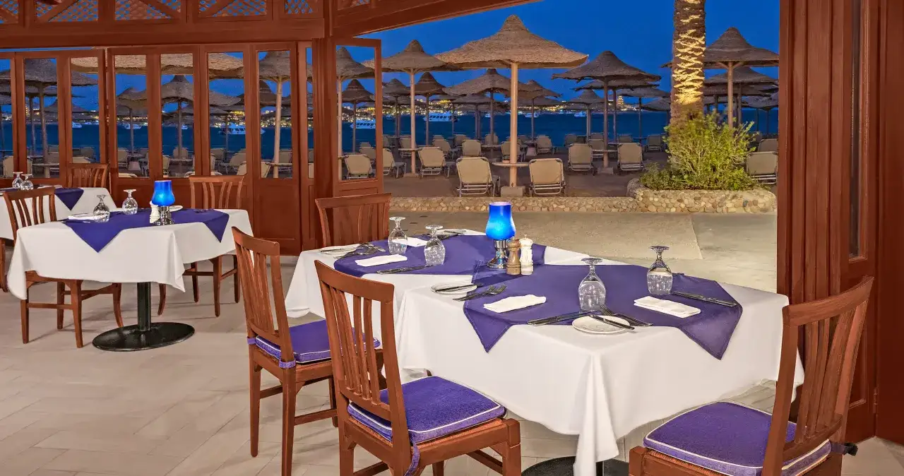 Elegancka restauracja nad morzem w Egipcie, gotowa na wieczór. Stoły nakryte białymi obrusami, fioletowe serwetki, a w tle plaża z parasolami. Pomyśl o napiwkach w Egipcie, by docenić obsługę.