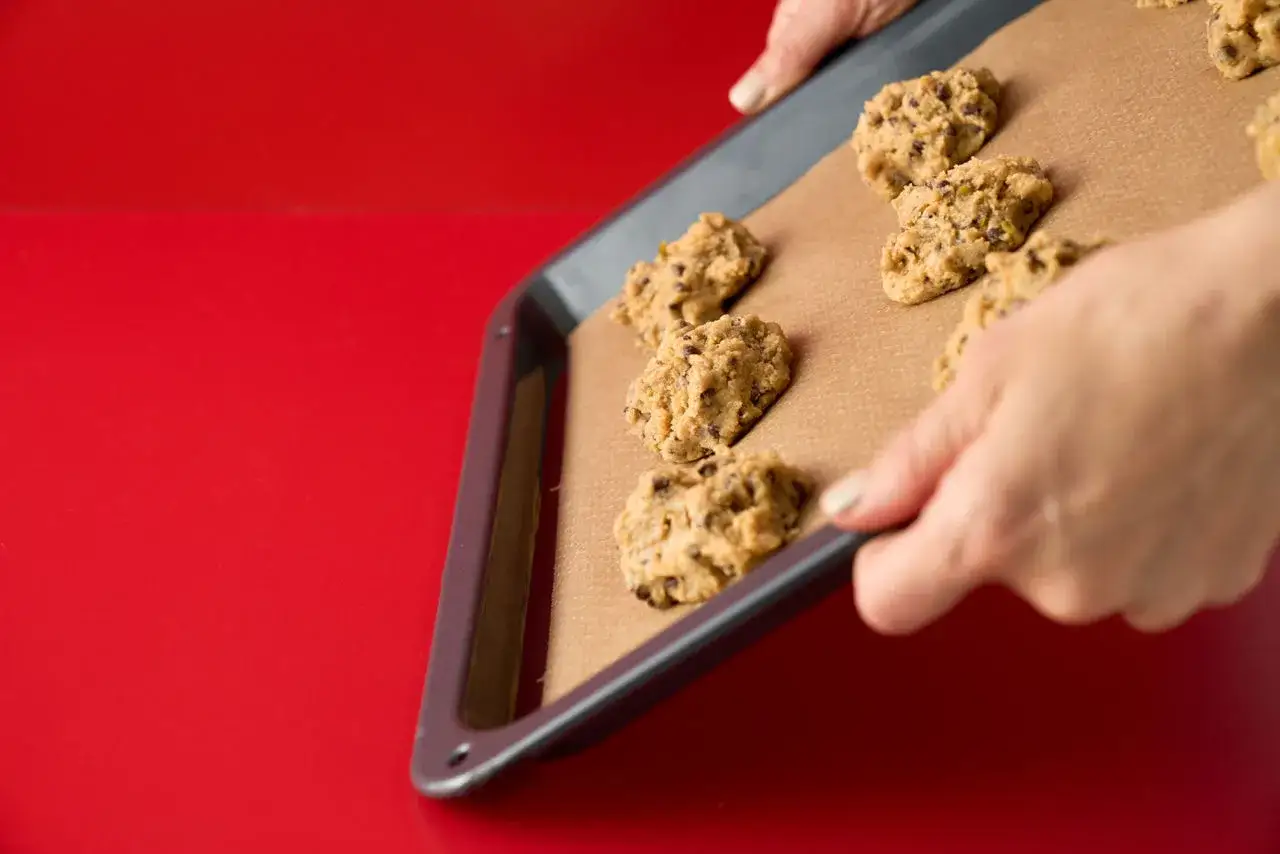 H&auml;nde halten ein Backblech mit frisch geformten Chocolate Chip Cookies auf Backpapier. Man muss Backpapier nicht einfetten, damit die Kekse nicht kleben bleiben.