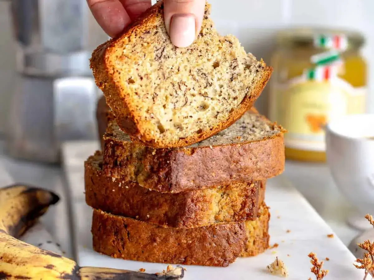 Una mano solleva una fetta di banana bread light, impilato su altre tre fette. Sullo sfondo, un barattolo di miele e una tazza di caffè.