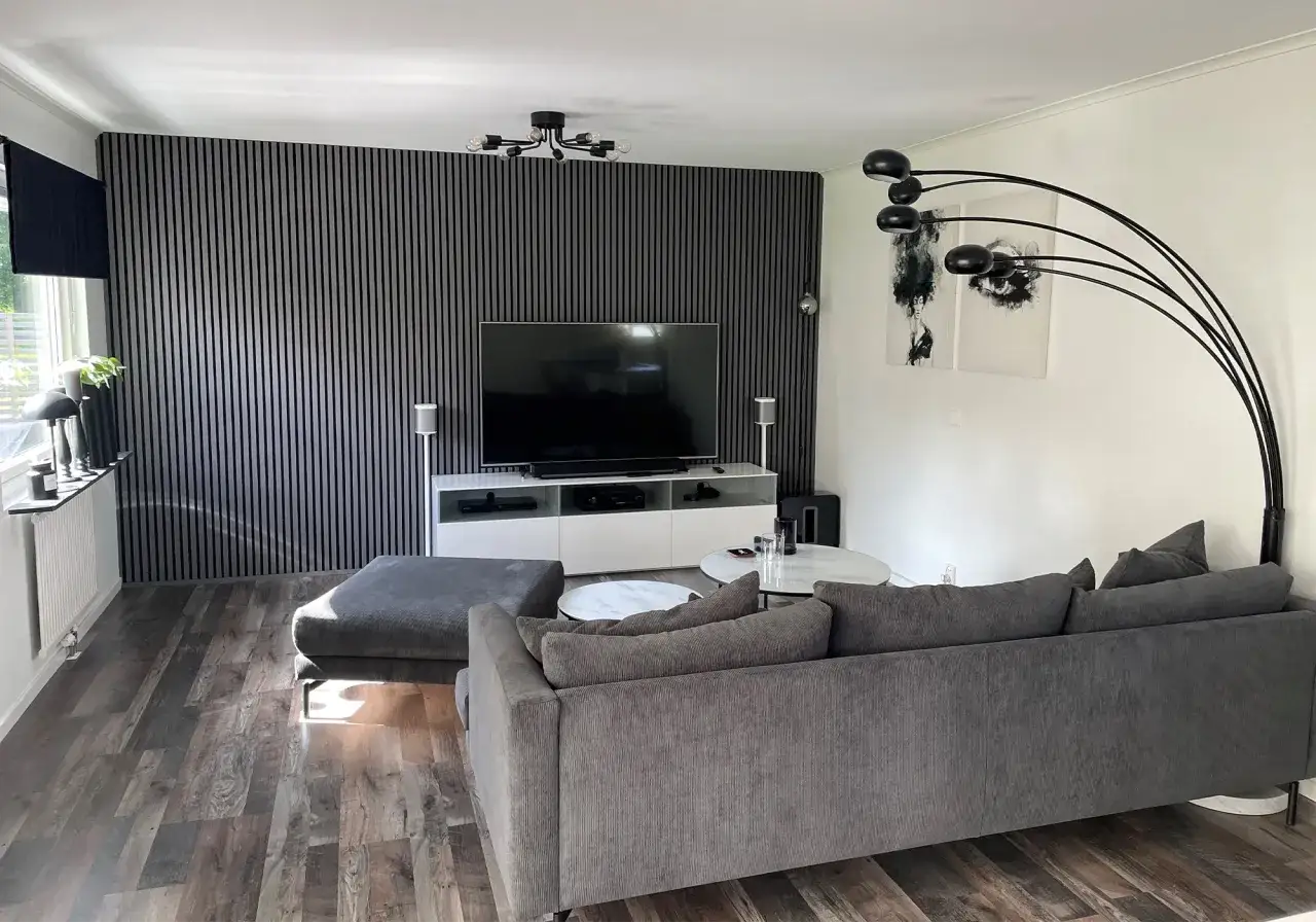 Modern inredning tv-rum med grå soffa, ribbad vägg och golv i träimitation. En stor TV står på ett vitt skåp.