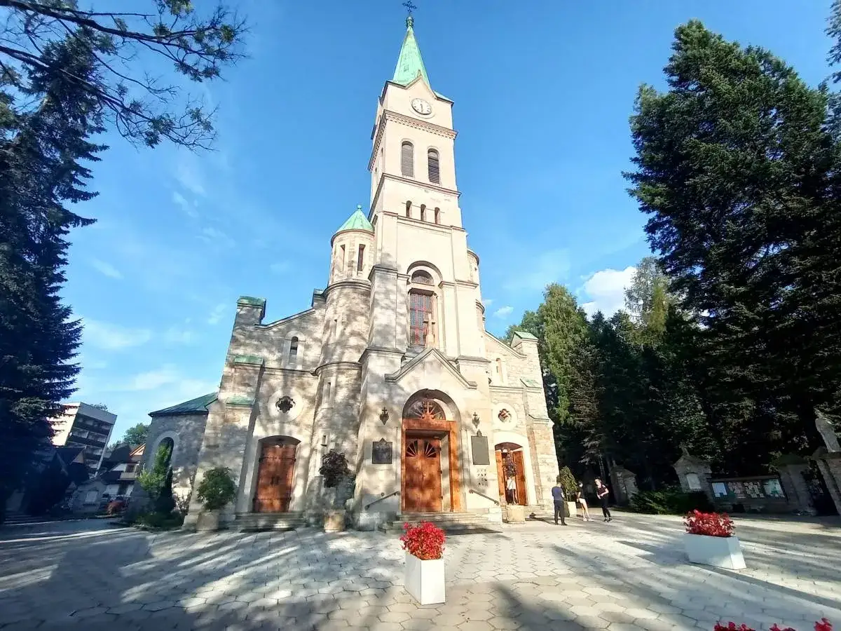 Słynny kościół w Zakopanem, z kamienną fasadą i zieloną wieżą, otoczony drzewami i błękitnym niebem.