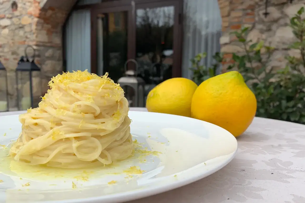Un piatto di spaghetti al limone cremosi, guarniti con scorza di limone grattugiata, accanto a due limoni freschi.