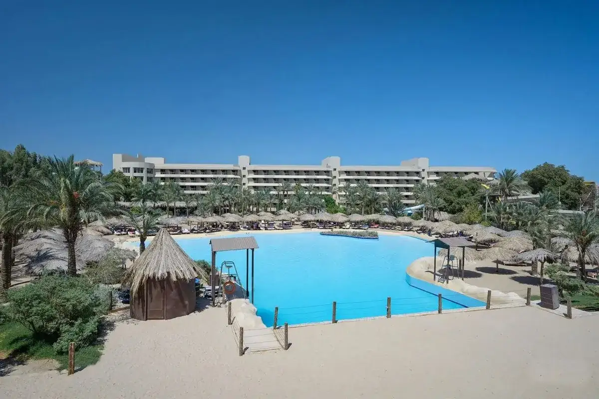 Duży basen z turkusową wodą, otoczony piaskiem, palmami i leżakami. W tle hotel, idealne miejsce na wakacje, o którym przeczytasz w sindbad club opinie.