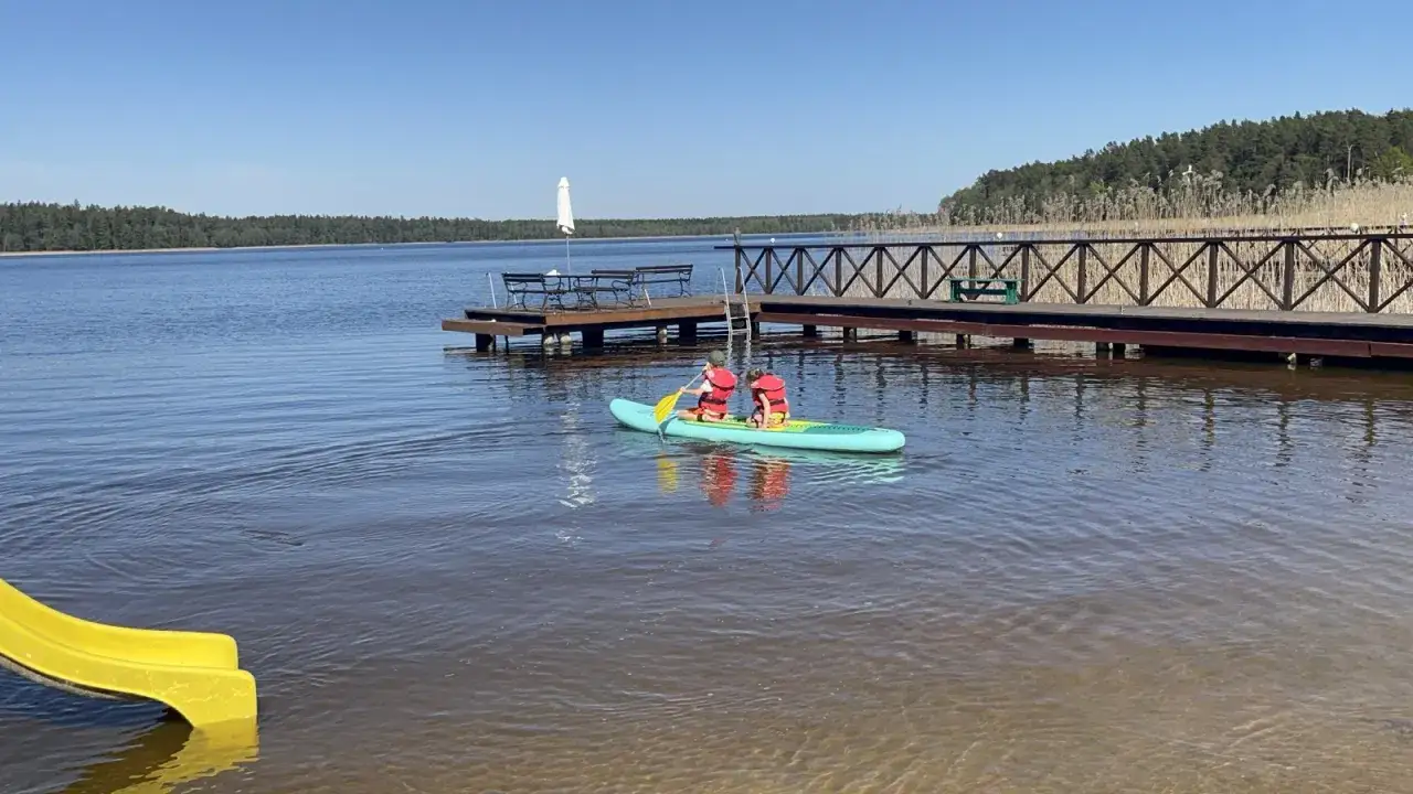 Dzieci na desce SUP płyną po spokojnym jeziorze w Augustowie. W tle drewniany pomost i las.
