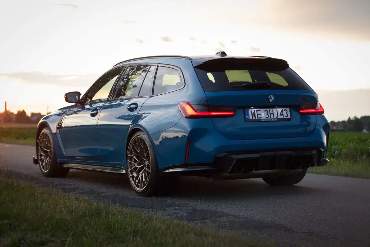 Niebieskie BMW M3 Touring z czterema końcówkami wydechu na tle zachodzącego słońca.