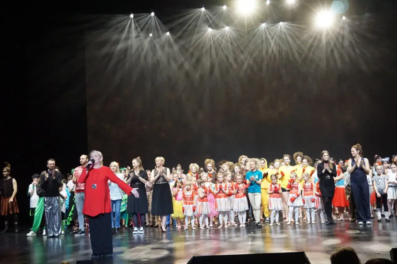 Wielka scena w Teatrze Gdynia, gdzie artyści i dzieci w barwnych strojach dziękują za oklaski po spektaklu.