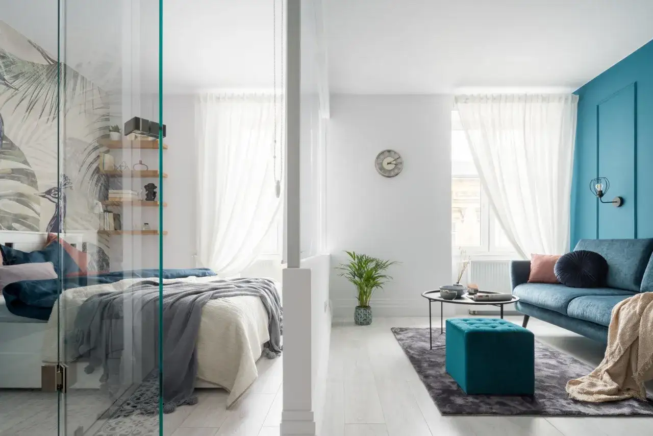 Nowoczesny salon z sypialnią. W sypialni łóżko z ozdobną tapetą, w salonie niebieska kanapa i turkusowa pufa.