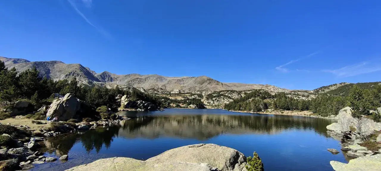 Lago sereno reflejando monta&ntilde;as y pinos. Un grupo descansa cerca de una roca, disfrutando de la paz del lugar. &iexcl;Pic carlit!