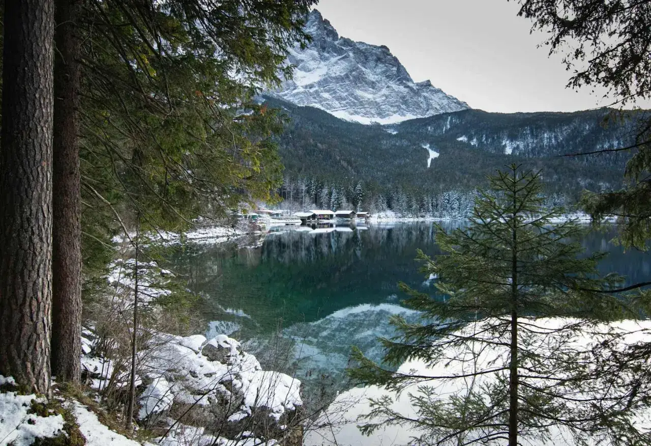 Eibsee winter: verschneite Berge spiegeln sich im klaren Wasser, Häuser am Ufer, Bäume im Vordergrund.