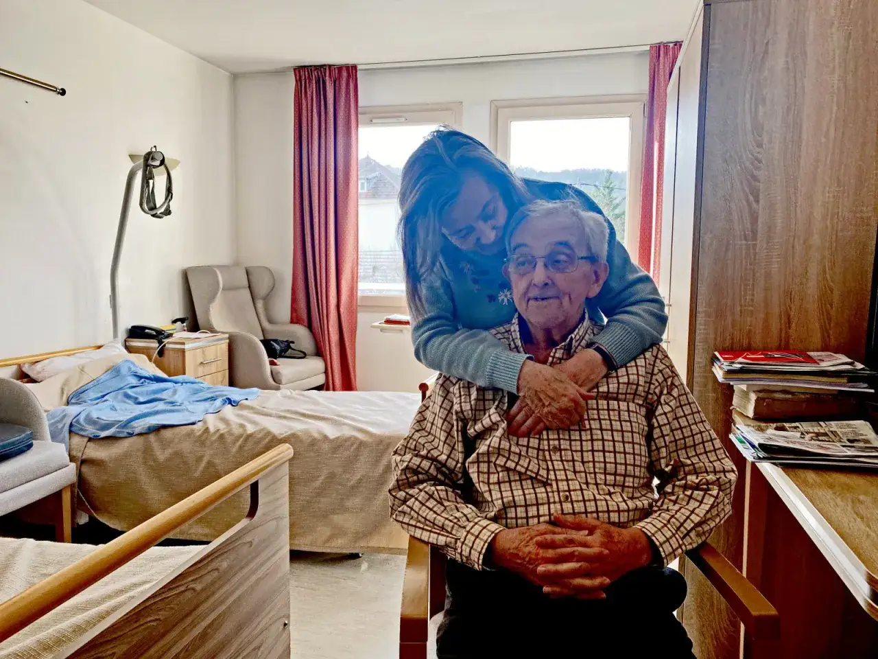 Une femme enlace tendrement un homme âgé dans une chambre d'EHPAD. Le tarif EHPAD pour un couple est une préoccupation, mais l'amour familial est inestimable.