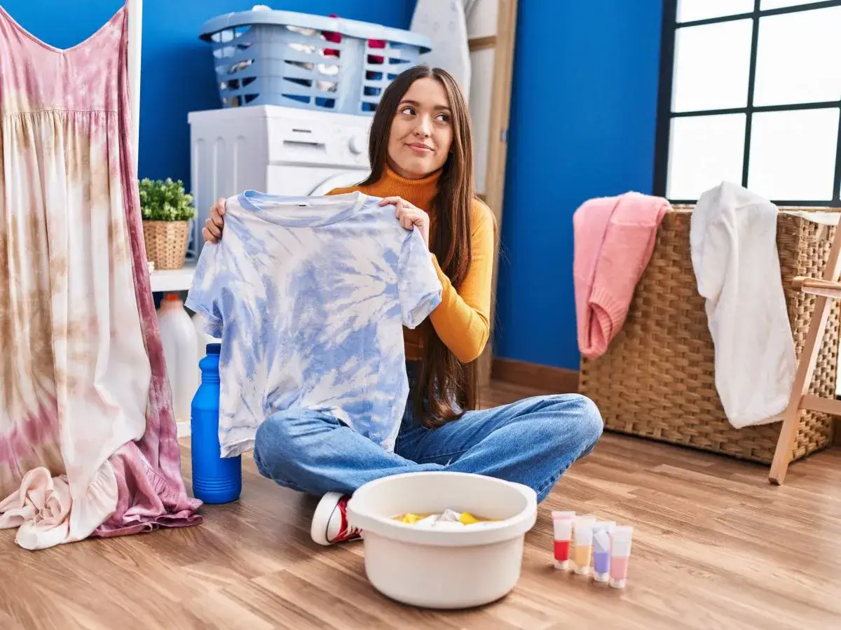 Kobieta z koszulą w stylu tie-dye zastanawia się nad farbowaniem ubrań. Obok miska z barwnikami i pralka.