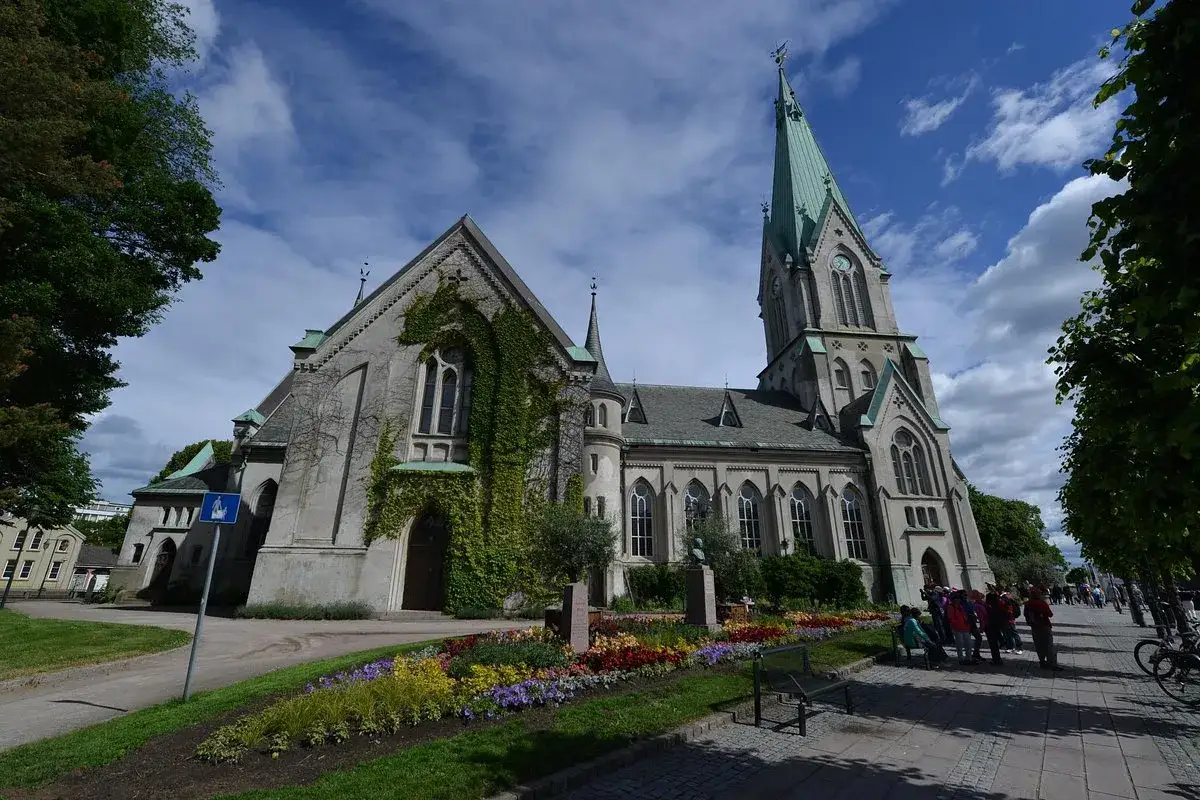 Imponerande kyrka med torn och grönt tak i Kristiansand, Norge. Blommor och träd pryder omgivningen.