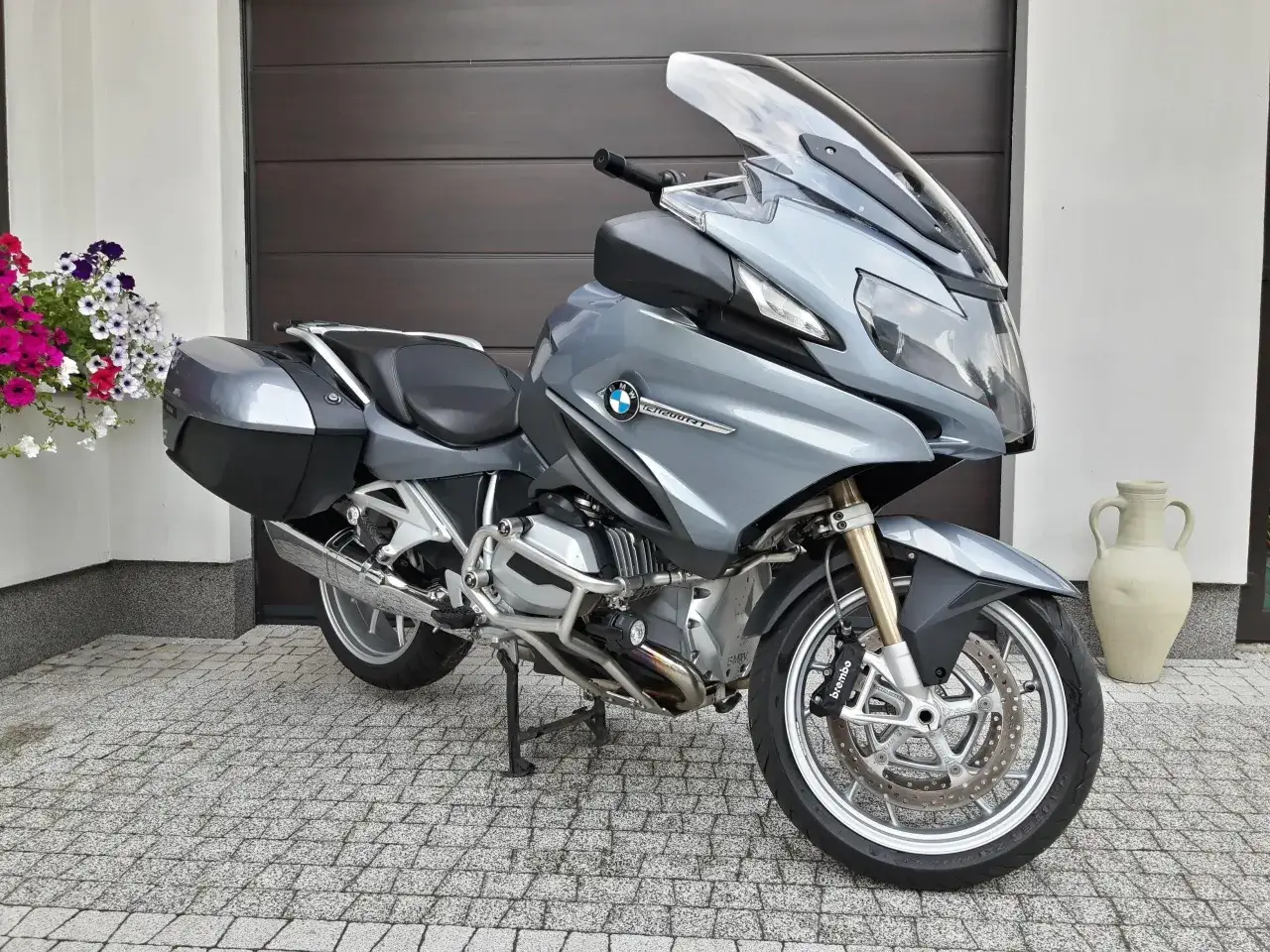 Szary motocykl BMW R 1200 RT z kuframi, zaparkowany przed garażem, obok donicy z kwiatami.