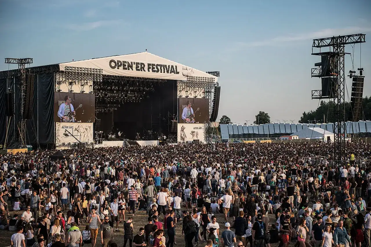 Tłum ludzi na festiwalu Open'er, przed wielką sceną z ekranami. W tle widać namioty i konstrukcje.