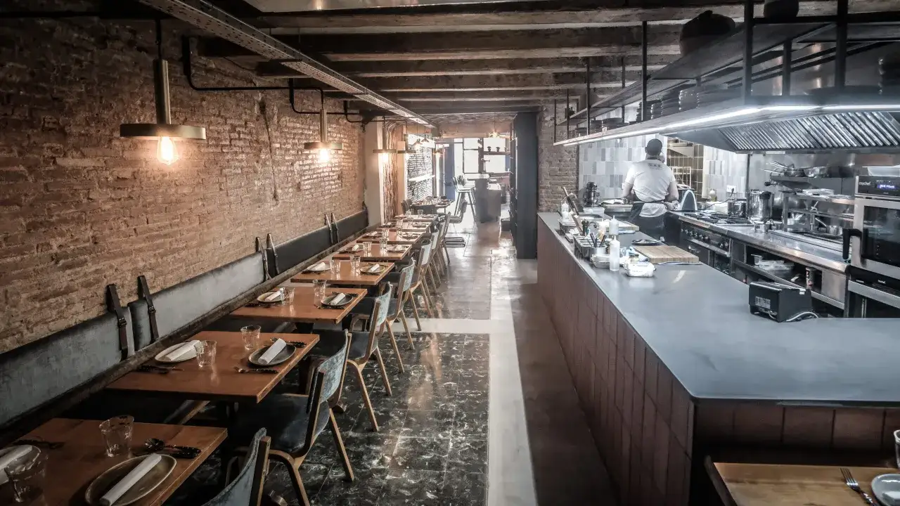 Interior de una taberna oeste con mesas de madera, asientos de terciopelo y una cocina abierta.