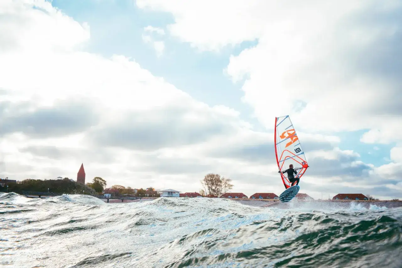 Ein Windsurfer gleitet über die Wellen, das bunte Segel im Wind. Die Küstenlinie mit Häusern und einem Turm im Hintergrund. Ein toller Tag zum pepelow surfen.