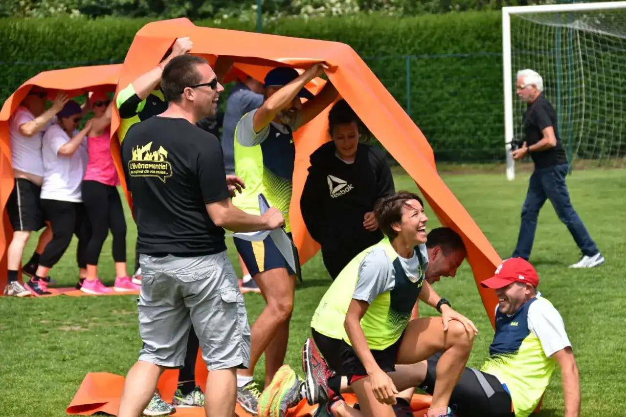 Des participants hilares lors d'un jeu de **team building jeux olympiques** sous un grand tissu orange.