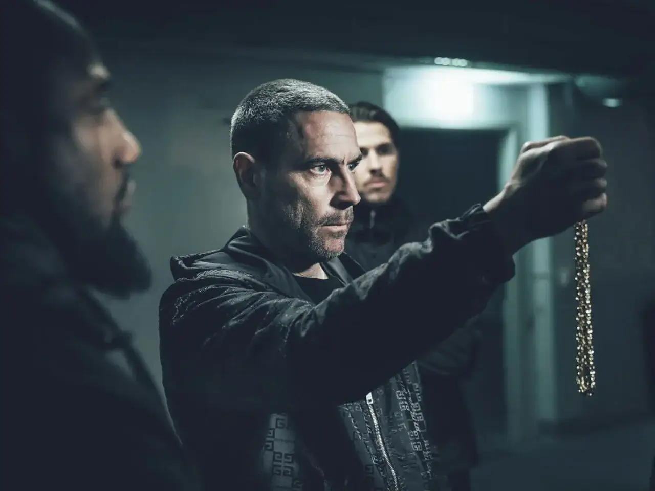 Sebastian Stakset film: en man h&aring;ller upp ett guldkedja, tv&aring; andra m&auml;n syns i bakgrunden.