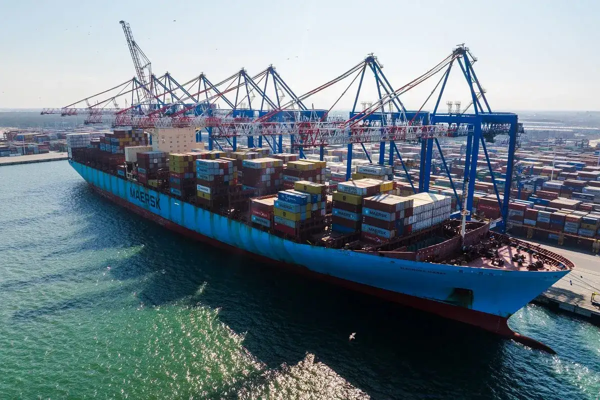 Ogromny kontenerowiec Maersk z pełnym ładunkiem przycumowany w porcie. W tle widać dźwigi i rzędy kontenerów. Rufa statku jest widoczna z boku.