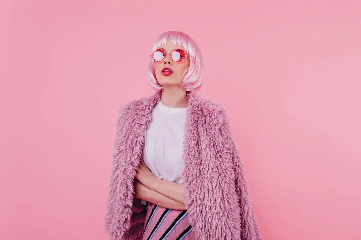 Dziewczyna w różowej peruce i okularach, w puszystym różowym płaszczu i spódnicy w paski. Inspiracja barbie stylizacje.