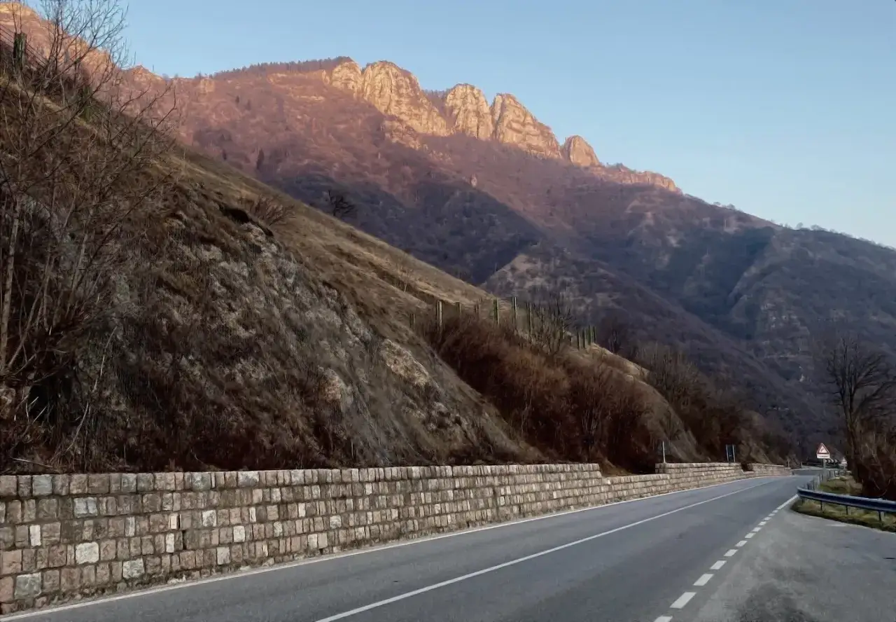 Strada del costo serpeggia tra montagne spoglie, illuminate da un sole dorato. Muri di pietra sorreggono il pendio.