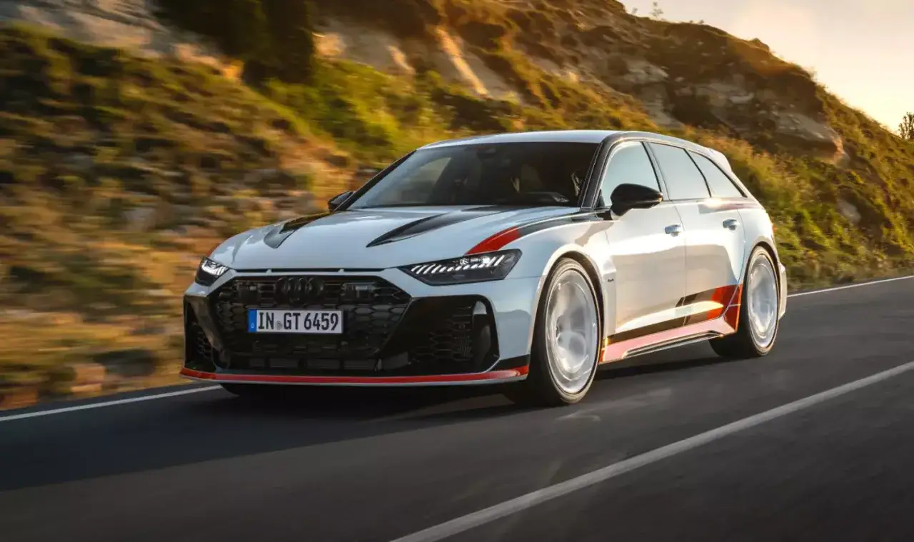 Białe Audi RS6 Avant z czerwonymi akcentami pędzi po krętej drodze wśród wzgórz o zachodzie słońca.