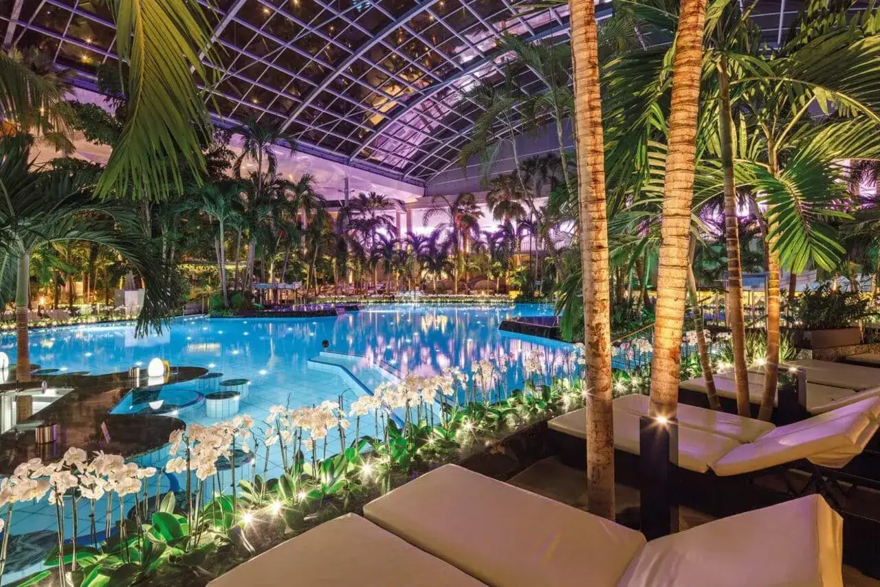 Tropische Palmen und ein leuchtend blauer Pool unter einem Glasdach – ein perfekter Urlaub in Deutschland. Entspannung pur mit Orchideen und Liegen.