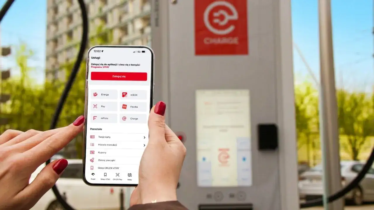 Kobieta używa aplikacji **Energa** na telefonie, by zalogować się do programu VITAY. W tle widać ładowarkę do samochodów elektrycznych.