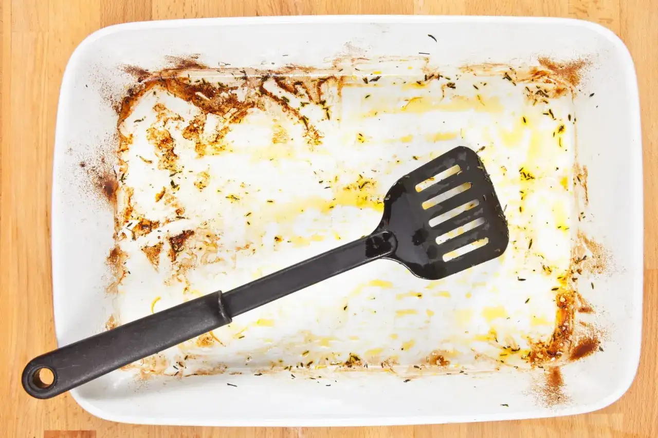 Une spatule noire repose dans un plat à four blanc sale, couvert de résidus brûlés et de graisse. Il est temps de nettoyer ce plat de four brûlé.